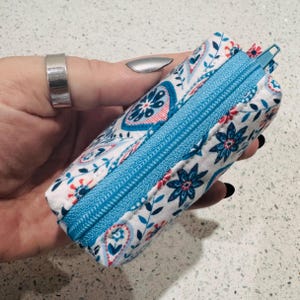 Puede incluir: Una bolsa rectangular con cremallera, con fondo blanco y estampado floral azul y rojo. La cremallera es de color azul brillante. La bolsa se sostiene en una mano.