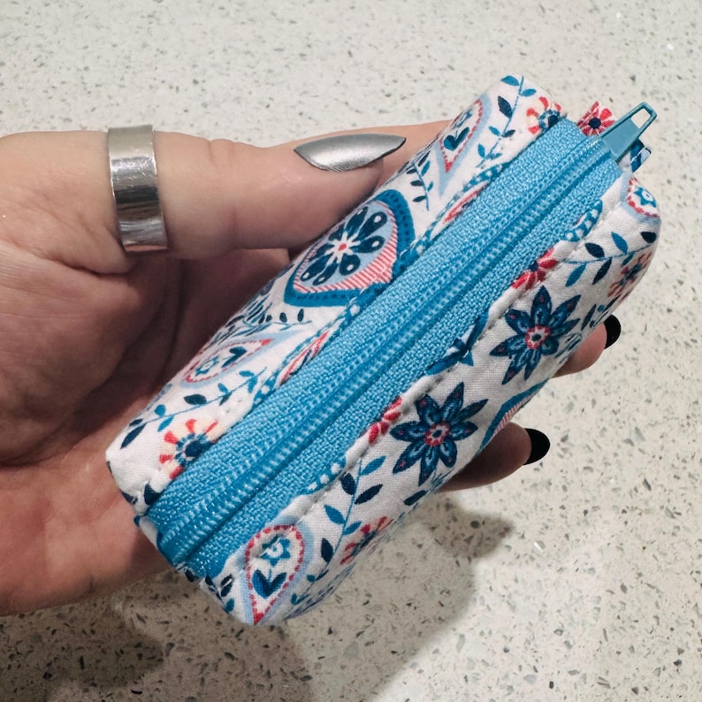 Puede incluir: Una bolsa rectangular con cremallera, con fondo blanco y estampado floral azul y rojo. La cremallera es de color azul brillante. La bolsa se sostiene en una mano.