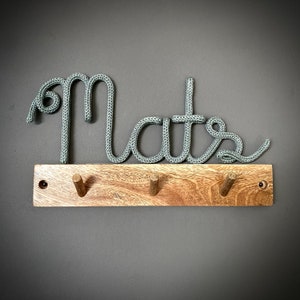 Könnte beinhalten: Ein Holz-Kleiderständer mit drei Haken, auf dem das Wort "Mates" in grauem Seil geschrieben ist.