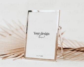 May include: A silver picture frame with a white mat and a white background. The frame is standing on a table with a dried palm leaf in the background. The text "Your design here!" is written on the white background. The text "5 x 7 CARD" is written in the bottom right corner of the white background.