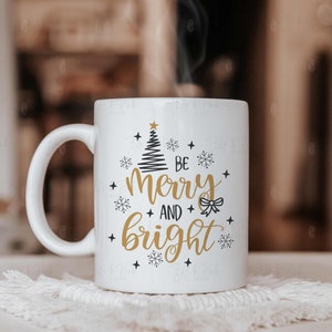 May include: White ceramic mug with a handle, featuring the text "Be Merry and Bright" in gold and black, along with a Christmas tree and snowflake design. The mug is placed on a white, textured coaster.