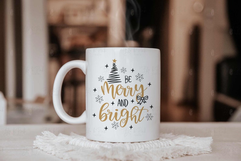 May include: White ceramic mug with a handle, featuring the text "Be Merry and Bright" in gold and black, along with a Christmas tree and snowflake design. The mug is placed on a white, textured coaster.
