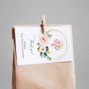 May include: Thank you!  A white card with gold lettering and a pink floral design with a chalice and host symbol. The card is attached to a brown paper bag with a wooden clothespin. The text on the card reads "Thank you! Alissa's First Holy Communion".