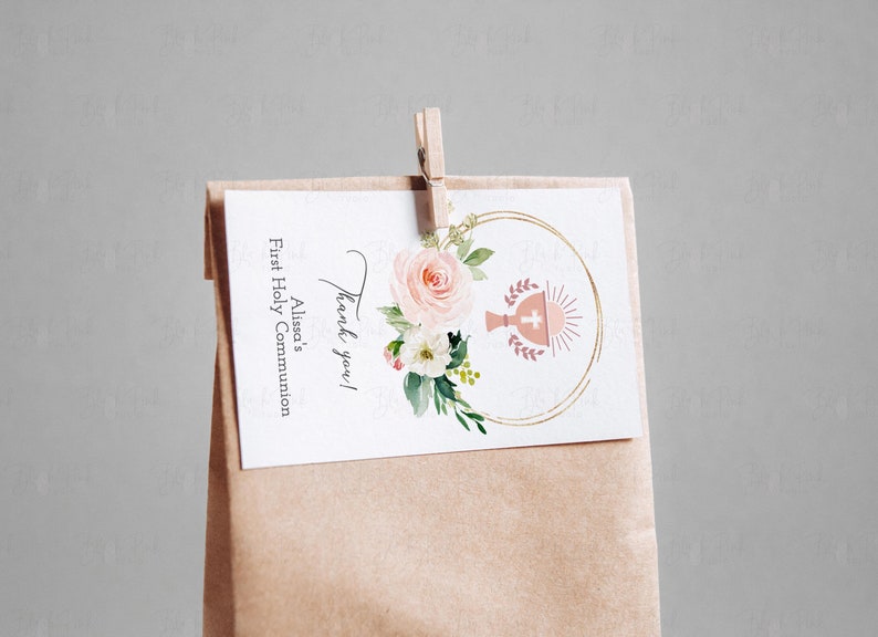 May include: Thank you!  A white card with gold lettering and a pink floral design with a chalice and host symbol. The card is attached to a brown paper bag with a wooden clothespin. The text on the card reads "Thank you! Alissa's First Holy Communion".