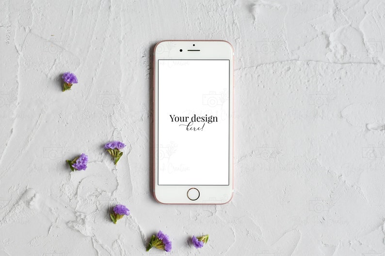 May include: A white smartphone with a rose gold frame and a blank screen. The text "Your design here!" is displayed on the screen. The phone is lying flat on a white textured surface with small purple flowers scattered around it.