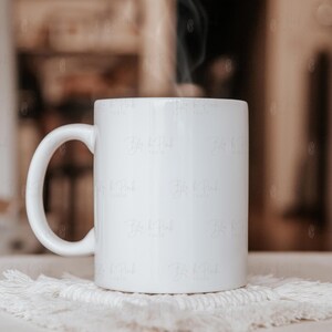 May include: A white ceramic mug with a curved handle sits on a white, textured coaster. Steam rises from the mug, suggesting a hot beverage. The background is softly blurred, with neutral tones.