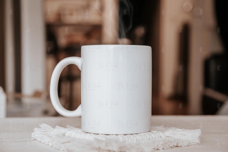 May include: A white ceramic mug with a curved handle sits on a white, textured coaster. Steam rises from the mug, suggesting a hot beverage. The background is softly blurred, with neutral tones.