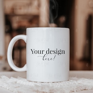May include: A white ceramic mug with a curved handle, resting on a white, textured coaster. The mug has the text "Your design here!" in black script. Steam rises from the mug, suggesting a warm beverage.