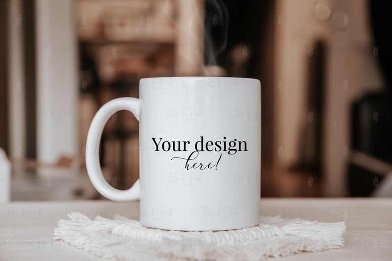 May include: A white ceramic mug with a curved handle, resting on a white, textured coaster. The mug has the text "Your design here!" in black script. Steam rises from the mug, suggesting a warm beverage.