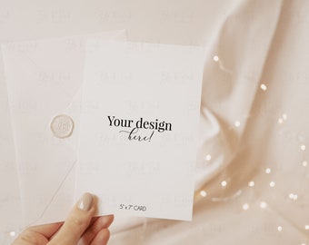 May include: A white card with the text "Your design here!" and "5 x 7 CARD" printed on it. The card is being held by a hand with light pink nail polish. The card is on top of a white envelope with a wax seal. The background is a light beige fabric with white fairy lights.