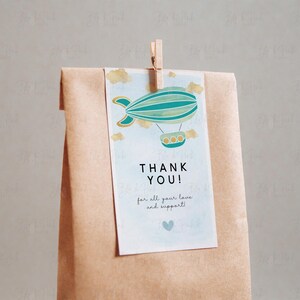 Puede incluir: Una bolsa de papel marr&oacute;n con una etiqueta de agradecimiento adjunta. La etiqueta presenta una aeronave azul y verde con detalles dorados y el texto "&iexcl;Gracias! por todo tu amor y apoyo!"