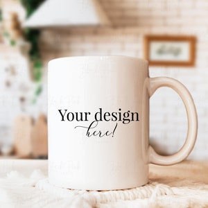 May include: A white ceramic mug with a curved handle, resting on a white textured surface. The mug features the text "Your design here!" in black script. The background is blurred, suggesting a kitchen setting.