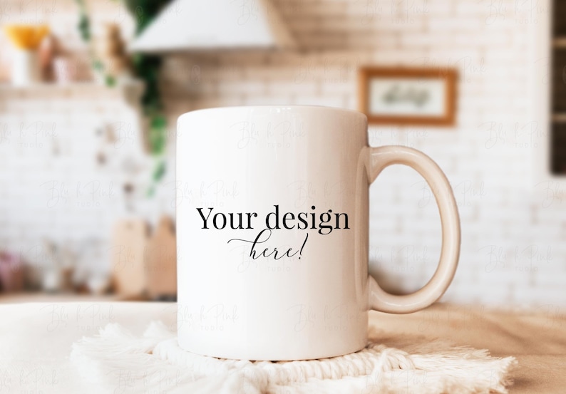 May include: A white ceramic mug with a curved handle, resting on a white textured surface. The mug features the text "Your design here!" in black script. The background is blurred, suggesting a kitchen setting.