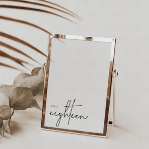May include: A silver-framed table number card with the word "eighteen" in elegant script. The card is propped up on a stand, with a neutral background and decorative foliage.