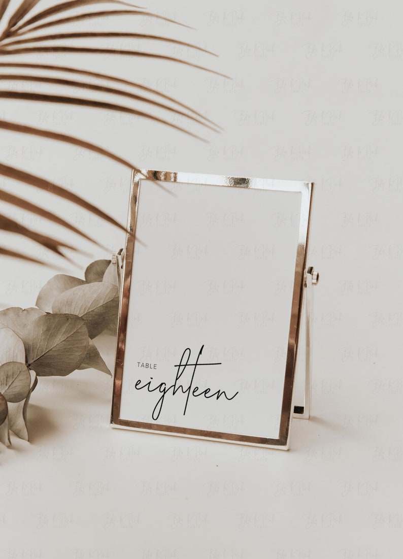 May include: A silver-framed table number card with the word "eighteen" in elegant script. The card is propped up on a stand, with a neutral background and decorative foliage.