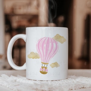 May include: White ceramic mug featuring a pink and white hot air balloon design with gold accents and yellow clouds. The mug has a curved handle and is sitting on a white, textured coaster. Steam rises from the mug.