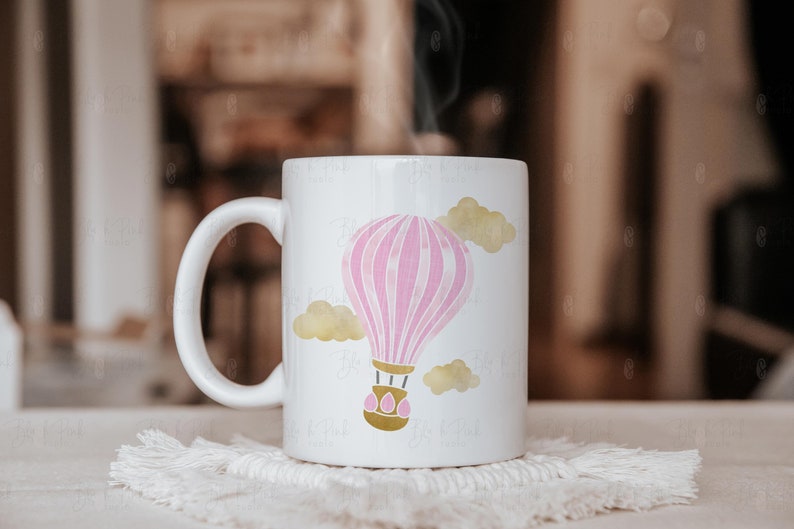 May include: White ceramic mug featuring a pink and white hot air balloon design with gold accents and yellow clouds. The mug has a curved handle and is sitting on a white, textured coaster. Steam rises from the mug.