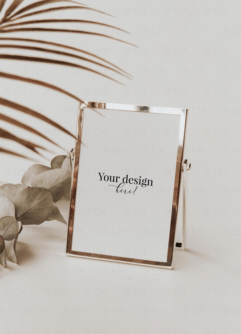 May include: A silver-framed photo or art display with the text "Your design here!" on a white background. The frame is rectangular and stands upright. Dried leaves and a palm frond are in the background, creating a neutral, minimalist aesthetic.