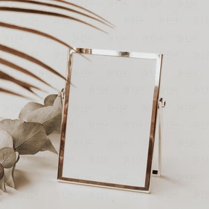 May include: A silver-framed tabletop mirror with a white interior, angled slightly. The mirror is supported by a hinged stand. Dried leaves and a palm frond are in the background, creating a neutral, minimalist aesthetic.