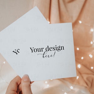 May include: Two white square cards with the text "Your design here!" are held in a hand. The cards are angled, with the text in a stylish font. The background is a soft peach fabric with fairy lights.