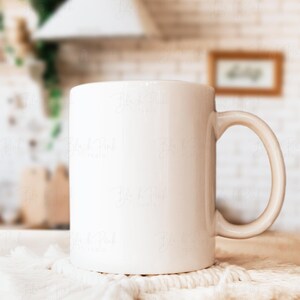 May include: A white ceramic mug with a curved handle sits on a white macrame coaster. The mug is cylindrical and appears to be empty. The background is a blurred kitchen scene with a white brick wall and wooden accents.