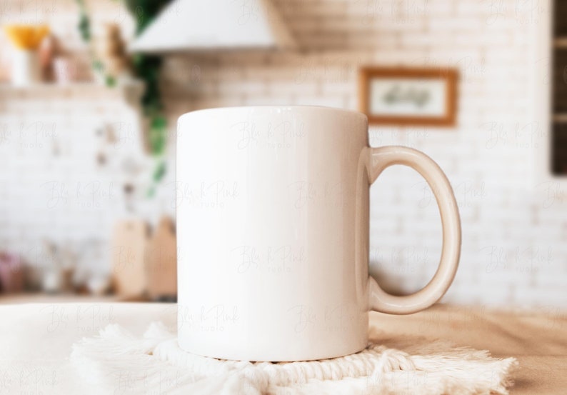 May include: A white ceramic mug with a curved handle sits on a white macrame coaster. The mug is cylindrical and appears to be empty. The background is a blurred kitchen scene with a white brick wall and wooden accents.