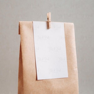 Puede incluir: Una bolsa de papel marr&oacute;n con una tarjeta blanca unida a ella con una pinza de madera. La tarjeta est&aacute; en blanco.