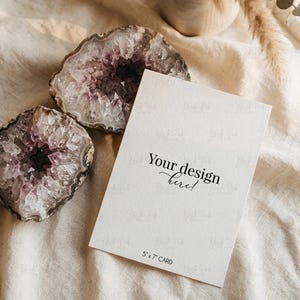 May include: A white 5x7 inch rectangular card with the black printed text Your design here! positioned next to two rough-edged amethyst geode halves featuring purple crystalline centers and grey outer rock layers on a textured cream fabric.