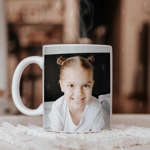 May include: White ceramic mug featuring a smiling child with two buns. The image on the mug is a close-up portrait of the child, wearing a white shirt. The mug is sitting on a white, textured coaster.
