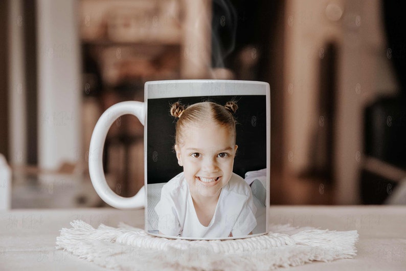 May include: White ceramic mug featuring a smiling child with two buns. The image on the mug is a close-up portrait of the child, wearing a white shirt. The mug is sitting on a white, textured coaster.