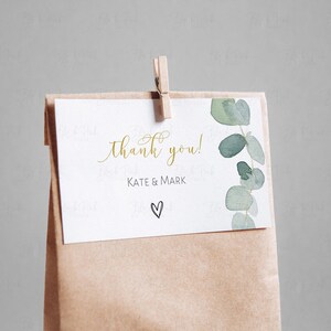 May include: A white thank you card with gold lettering that says "thank you!" and "Kate & Mark" with a small hand-drawn heart. The card is attached to a brown paper bag with a wooden clothespin.