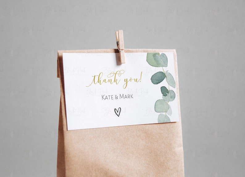 May include: A white thank you card with gold lettering that says "thank you!" and "Kate & Mark" with a small hand-drawn heart. The card is attached to a brown paper bag with a wooden clothespin.