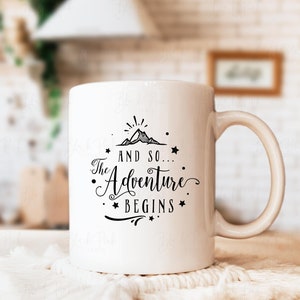 May include: White ceramic mug with a black graphic that reads "And so... The Adventure Begins" with a mountain and star design. The mug has a curved handle and is set on a white surface.