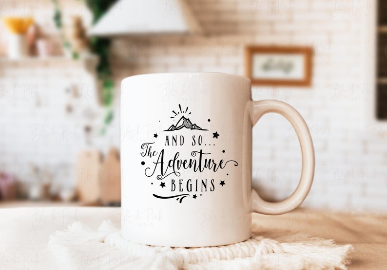 May include: White ceramic mug with a black graphic that reads "And so... The Adventure Begins" with a mountain and star design. The mug has a curved handle and is set on a white surface.