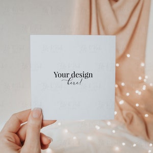 May include: A square white card held by a hand, with the text "Your design here!" in elegant script. The background features a soft, draped fabric in a peach hue, with twinkling fairy lights adding a warm glow.
