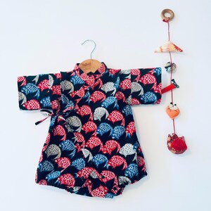 May include: A baby's kimono-style romper with a vibrant fish pattern in red, blue, and gray on a black background. The romper is displayed on a wooden hanger. A decorative mobile with fabric ornaments hangs nearby.