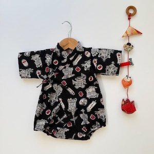May include: Black baby kimono-style romper with a repeating pattern of Japanese-style illustrations in white and red. The romper is hanging on a wooden hanger. A decorative mobile with wooden and fabric elements hangs to the right.