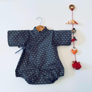 Könnte beinhalten: Ein marineblauer Baby-Kimono mit einem geometrischen Sternmuster, der an einem Holzbügel hängt. Ein dekoratives Mobile mit Stoffornamenten in Rot, Orange und Blumenmustern hängt in der Nähe.