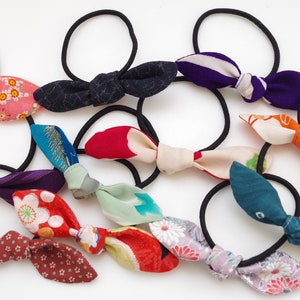 Può includere: Una collezione di elastici per capelli colorati con fiocchi di stoffa. I fiocchi sono realizzati con tessuti a motivi diversi, tra cui motivi floreali, geometrici e astratti. Gli elastici per capelli sono neri ed elastici.