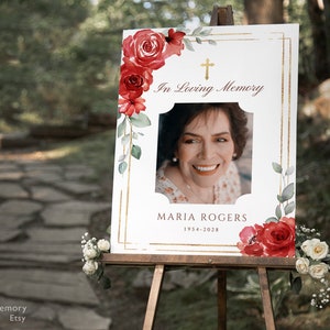 May include: A white memorial sign with a gold frame and red floral accents. The sign reads "In Loving Memory" and features a photo of a woman with the name "Maria Rogers" and the dates "1954-2028".