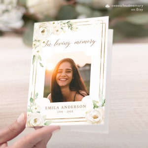 May include: White floral sympathy card with a gold frame and a photo of a smiling woman. The card reads "In loving memory" and "EMILA ANDERSON 1995 - 202X".