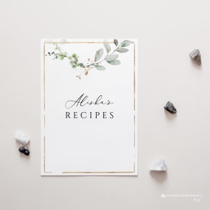 Op de afbeelding: Een witte kaart met een gouden frame en een groen en gouden bloemenontwerp. De tekst "Alisha's Recipes" is in zwart gedrukt.