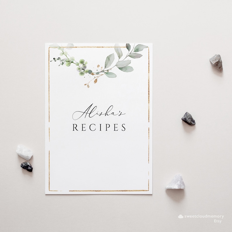 Op de afbeelding: Een witte kaart met een gouden frame en een groen en gouden bloemenontwerp. De tekst "Alisha's Recipes" is in zwart gedrukt.