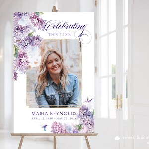 May include: A memorial sign with a watercolor floral design featuring purple and white flowers. The sign has a gold frame around a photo of a woman. The text reads "Celebrating the Life Maria Reynolds April 12, 1980 - May 20, 20XX".