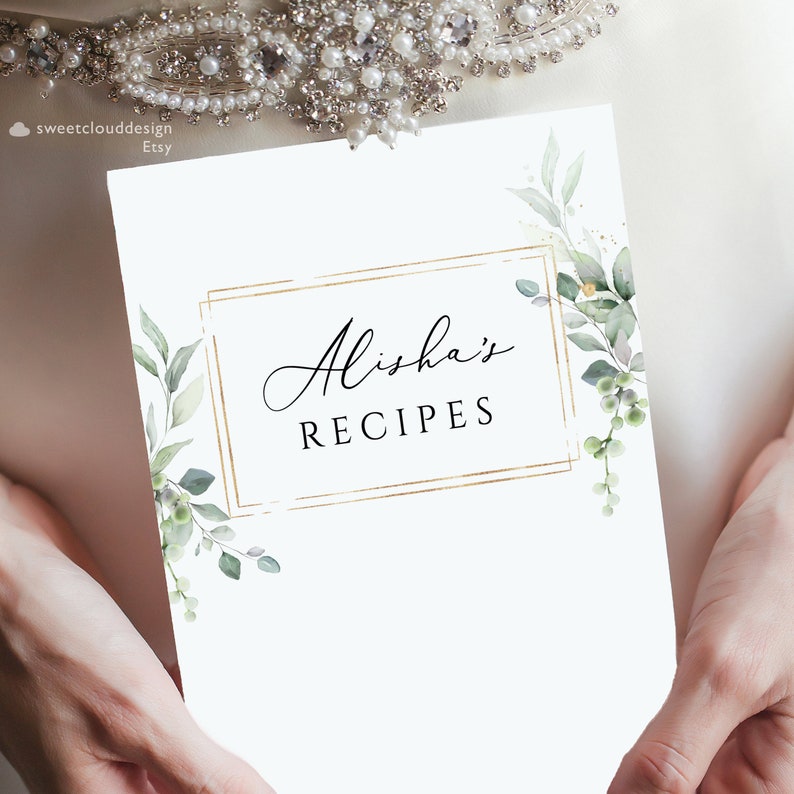 May include: A white recipe book cover with a gold frame and the text "Alisha's Recipes" in black cursive font. The cover is decorated with watercolor illustrations of green leaves and gold glitter.