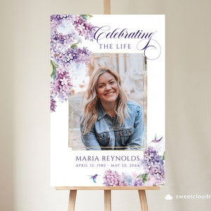 May include: A white memorial sign with a gold frame and a photo of a woman. The sign has a watercolor floral design with purple flowers and a hummingbird. The text reads "Celebrating the Life of Maria Reynolds, April 12, 1980 - May 20, 20XX".