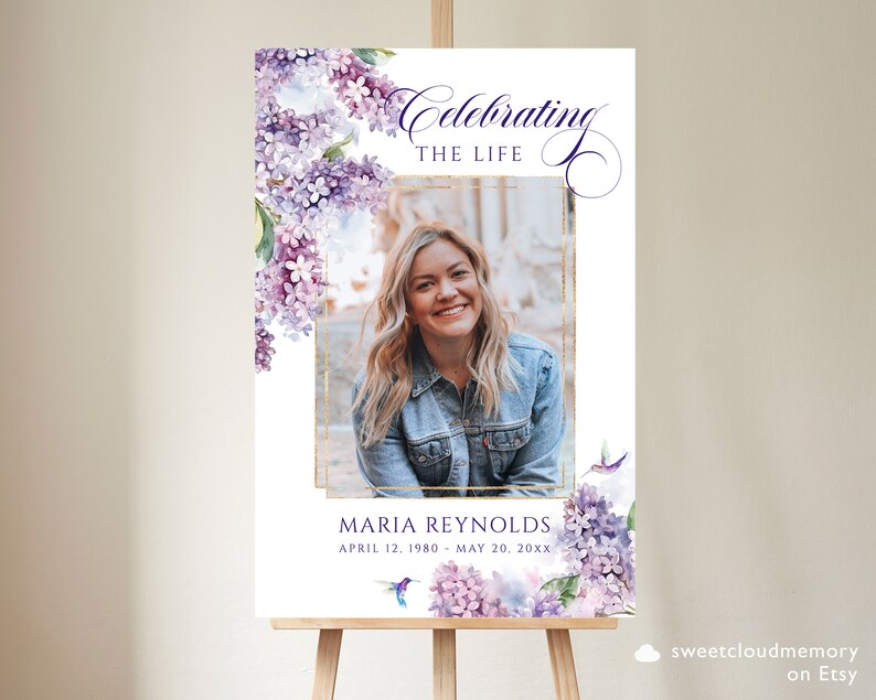 May include: A white memorial sign with a gold frame and a photo of a woman. The sign has a watercolor floral design with purple flowers and a hummingbird. The text reads "Celebrating the Life of Maria Reynolds, April 12, 1980 - May 20, 20XX".