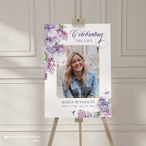 May include: A white memorial sign with a purple floral design and a gold frame around a photo of a woman. The text reads "Celebrating the Life of Maria Reynolds, April 12, 1980 - May 20, 20XX".