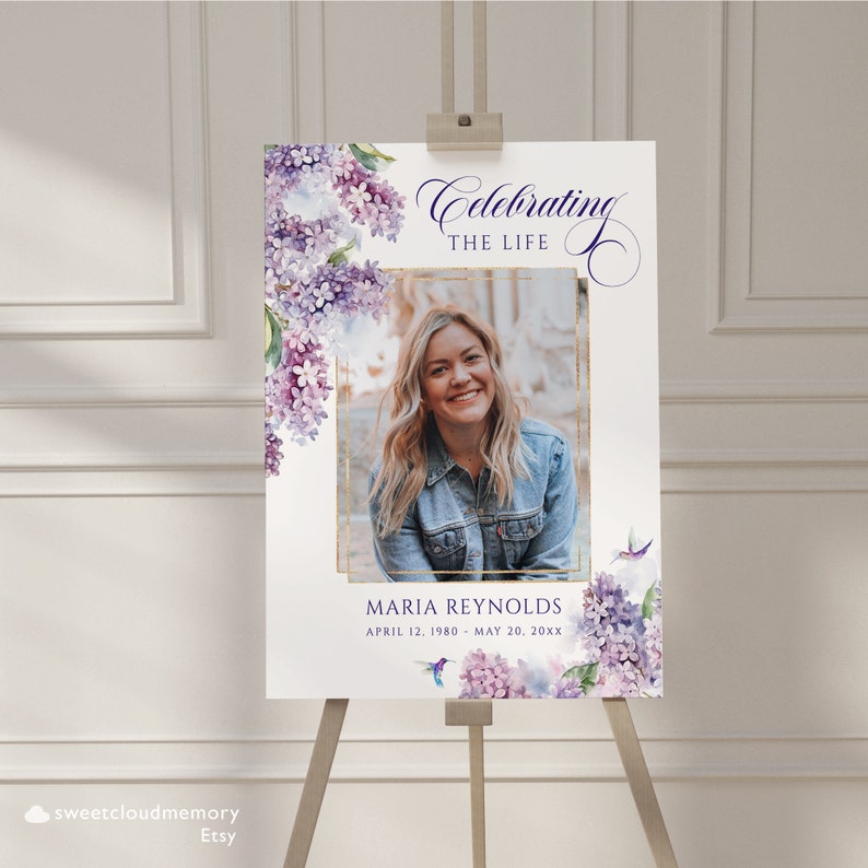 May include: A white memorial sign with a purple floral design and a gold frame around a photo of a woman. The text reads "Celebrating the Life of Maria Reynolds, April 12, 1980 - May 20, 20XX".