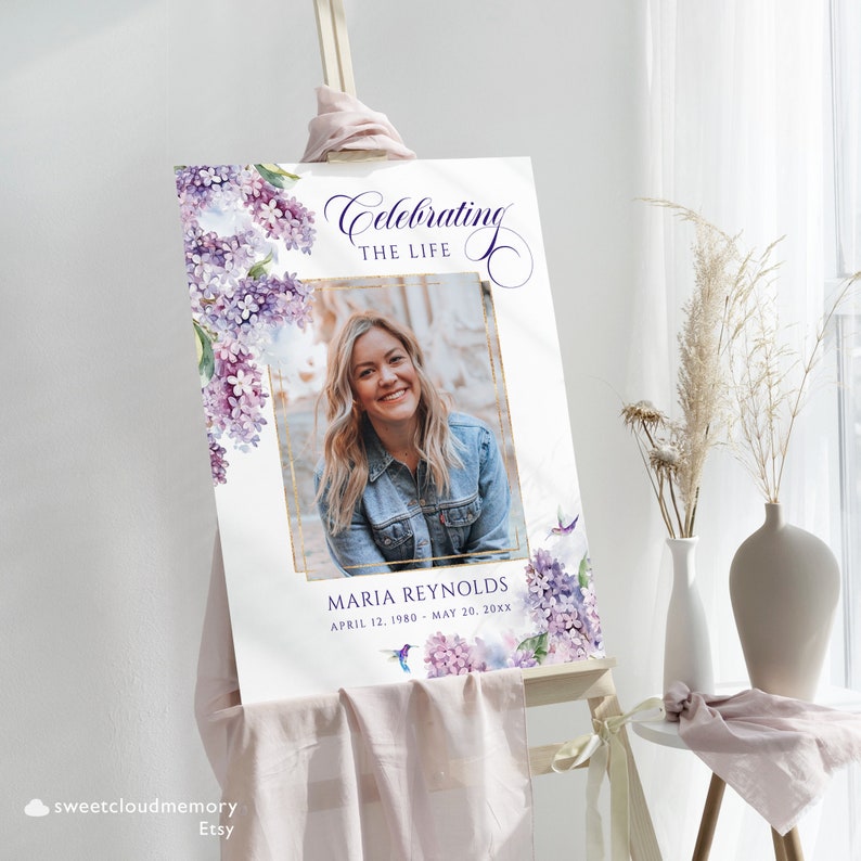 May include: A white memorial sign with a photo of a woman in a blue denim jacket. The sign is decorated with purple and blue watercolor flowers and the text "Celebrating the Life" and "Maria Reynolds April 12, 1980 - May 20, 20XX".